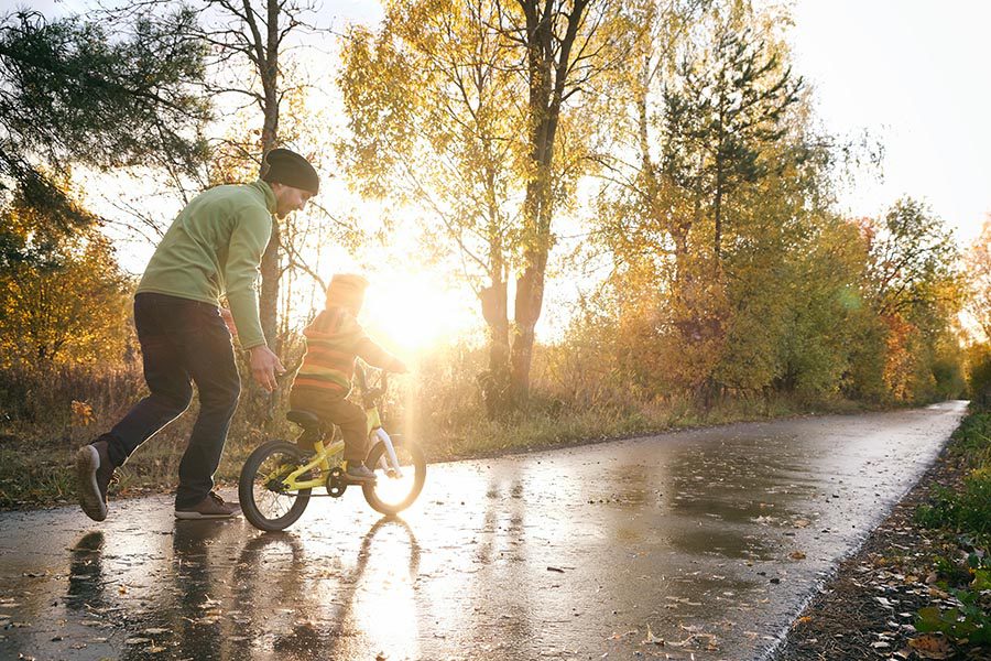 About Our Agency - Father and Young Son on a Wet Path in a Park, Father Teaching the Son How to Ride a Bike